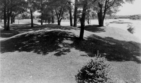 View of burial mounds in a park on the edge of a lake. Caption reads: "Indian Burial Grounds in Wakanda Park, Menomonie, Wis."