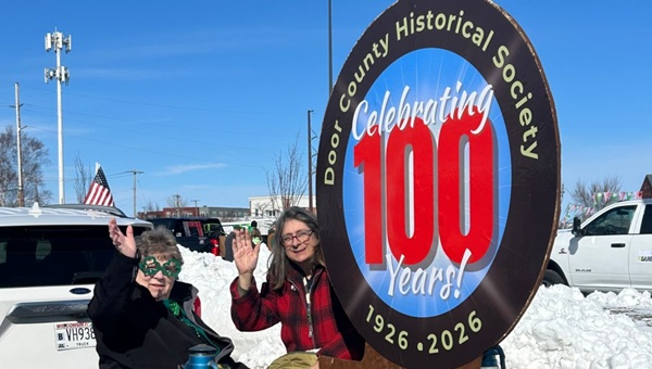 Two women sitting by a car and a 100th anniversary sign for the Door County Historical Society smile and wave