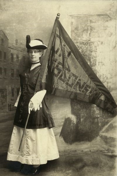 Portrait of Theodora Youmans (Mrs. Henry), President of the Wisconsin Woman Suffrage Association, holding a Wisconsin flag and standing in front of a painted backdrop