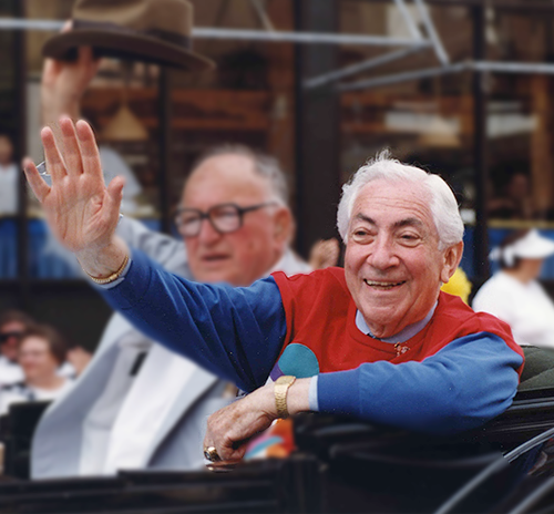 Ben Barkin rides in a vintage convertible waving happily to the crowd during a parade. His shirt is bright red and blue color block.