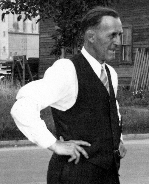 Billy Schultz wears a black and white suit with no jacket. His hands are on his hips and he squints with a slight grimace at something off camera. He's facing profile and to the right.