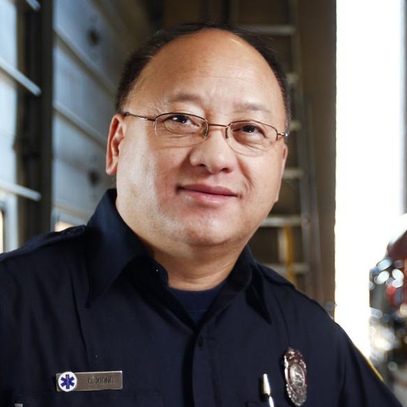 Headshot photo of Bouachao Xiong. Standing in font of a blurred background of the interior of a fire station, he's wearing his dark blue firefighter uniform with his hand resting on a nearby firetruck. He wears glass and smiles slightly at the camera.