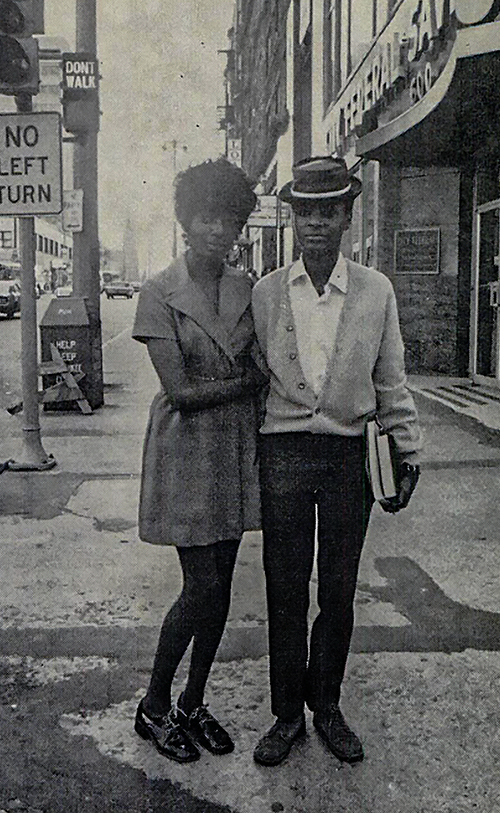 Manonia Evans (left) and Donna Burkett, 1971. Manonia Evans wears a wide labeled dress with short almost capped sleeves, her hair is curly and short, a small smile plays across her lips. Donna Burkett wearing a pork pie hat with a light ribbon, set at a jaunty angle and casual light suit. They stand close together with Evans' arm wrapped around one of Burkett's.