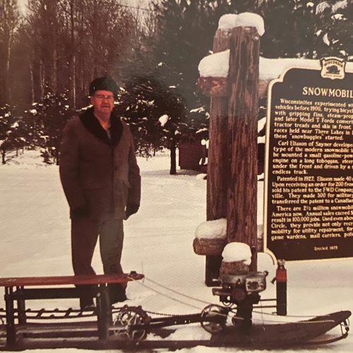 Carl Eliason, inventor of the modern day snowmobile, stands next a snowmobile and the historic marker in Sayner during winter with his fur lined winter clothes.