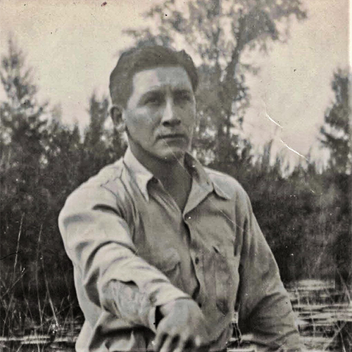 Jerome looking seriously off into the distance while in a canoe. Photograph was taken in 1942 on the Bad River Reservation, probably in the Kakagon sloughs.  He captioned it, The Hunter. You can see his rifle so we know he's out hunting. Based on that and how the rice beds behind him look, likely taken in autumn.