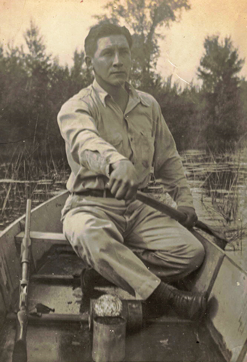 Jerome looking seriously off into the distance while in a canoe. Photograph was taken in 1942 on the Bad River Reservation, probably in the Kakagon sloughs.  He captioned it, The Hunter. You can see his rifle so we know he's out hunting. Based on that and how the rice beds behind him look, likely taken in autumn.
