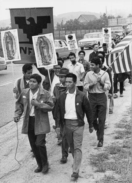 Madison on Highway 21 to petition lawmakers to hold farms and food industry corporations accountable for better working conditions for migrant farm workers. Jesús Salas, in front leading the march, is holding a microphone. Next to him is Salvador Sanchez, also a march leader. Two men behind Salas carry images of Our Lady of Guadalupe. One man in glasses is carrying the National Farm Workers Association banner with the Aztec eagle symbol. To the right, a man is holding the American flag. With Father Garrigan, in the middle of the march, are two men holding signs that say 'Juntarnos para ser reconocidos. Hablar para ser oidos. La raza tiene causa