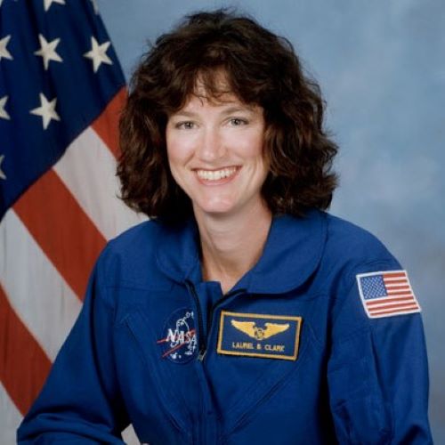 Laurel Clark in her astronaut uniform smiling at the camera with the american flag behind her.