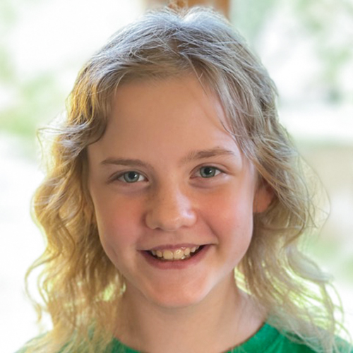 Liza, a young wavy haired blonde girl with blue eyes, smiles straight on towards the camera wearing a green t-shirt.