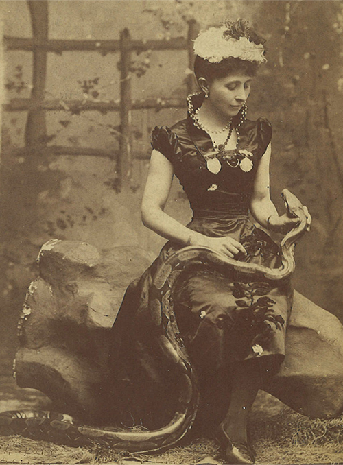 Lou Ringling, dressed in Edwardian period dark clothing, sits and looks comfortably at her giant python that is draped across her lap and onto the floor. This amazing woman also has a white feathered cap in her curly pinned up hair.