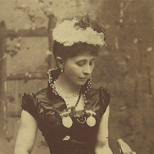 This amazing woman, dressed in Edwardian period dark clothing, sits and looks comfortably at her giant python that is draped across her lap and onto the floor. She has a feathered cap.