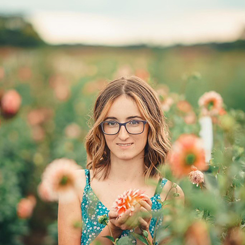 Madelyn, a short brown haired young woman wearing glasses, stands in a field of flowers wearing a teal floral tank dress. She holds one bloom and smiles slightly at the camera. 