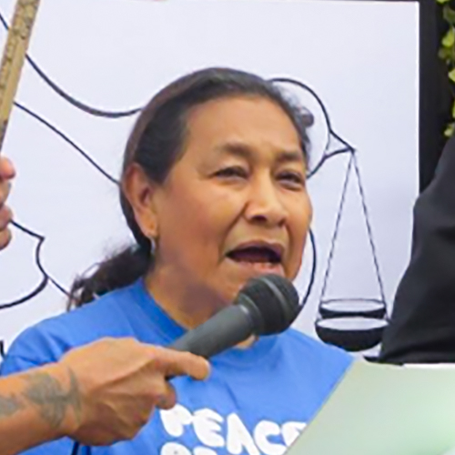 Maria Luisa Morales speaking at a demonstration for peace. She's wearing a blue shirt for the demonstration and her dark hair pulled back into a ponytail, scales of justice are visible behind her.