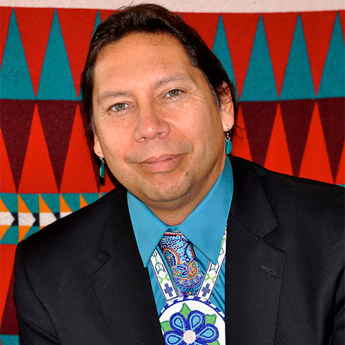 Marin Denning looking at the camera wearing a tradionally beaded neck piece over a paisley print tie and a teal shirt in a black suit jacket. The background is a brightly printed tapestry, with primarily triangle motifies.