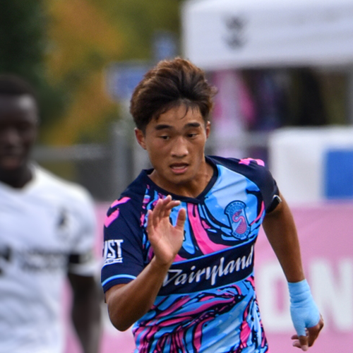 Michael Vang plays soccer, running hard and fast towards the camera eye on the ball out of frame, another play close behind over his shoulder.