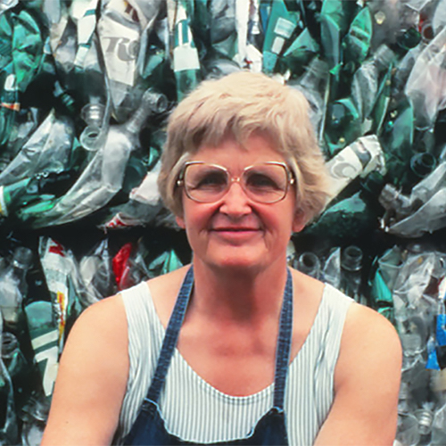 Longtime Sauk County resident Milly Zantow (1923-2014) sits in front of a wall of plastic, crushed into cubes ready to be recycled. She smiles slightly at the camera, wearing glasses, a white tank top, and a blue apron. 