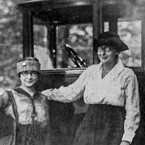 Miriam Frank and Charlotte Partridge stand in front of a car, smiling slightly in this informal portrait. They are both wearing long black skirts, and lighter colored blouses. Bespeckled with round frames and both wear hats.