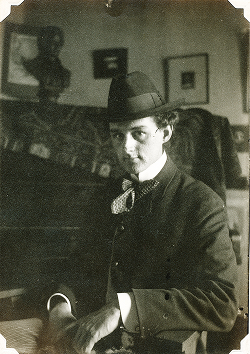 Photo Credit: Cooksville Archives and Collections Ralph Warner, looks almost temptingly at the camera, chin tilted down, hat on while sitting in a chair in a haphazardly decorated room.