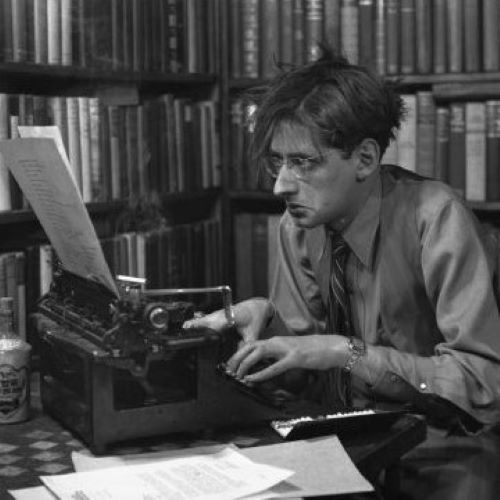 Robert Albert Bloch, author of Psycho, surrounded by tall bookshelves full of books, is sitting in front of a typewriter with an open cigarette case full of cigarettes, a bottle of Wing Fhung Hong, and a pile of papers.