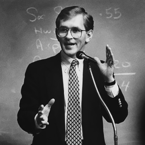 Steve Gunderson gestures animatedly in front of chalkboard with equations behind him. Wearing a suit and fun tie he seems excited and engaged.