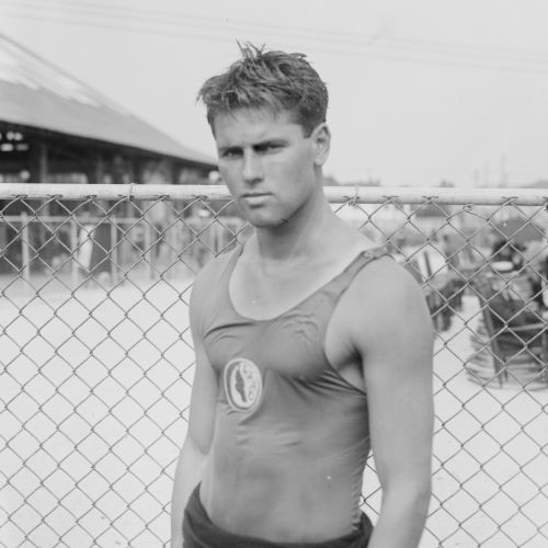 Tom Blake (shown in 1922) a handsome young man wears a swiming costume in front of a wire fence around a swimming pool. He looks steelily at the camera, a bit of a challenge in his eyes hair messy from swimming.