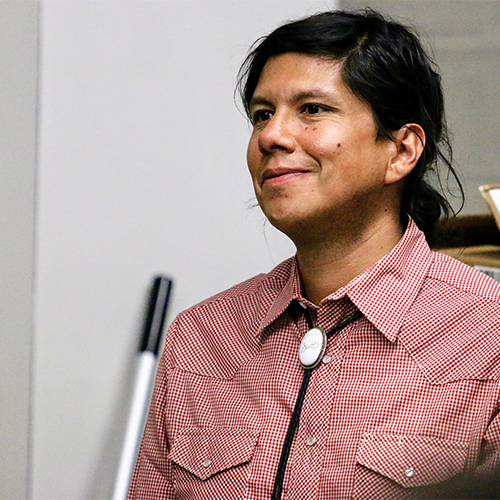 A Ho-Chunk man, long hair pulled back in a low pony tail, bolo tie, and red south west style shirt, smiles slightly as he sees the historic canoe for the first time.