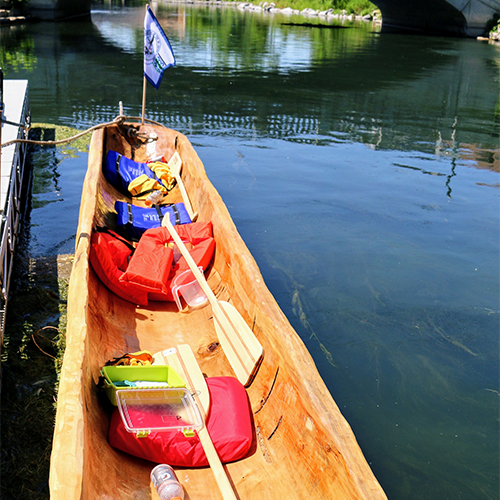 The canoe sits empty of people but filled with life vests and paddles on the river.