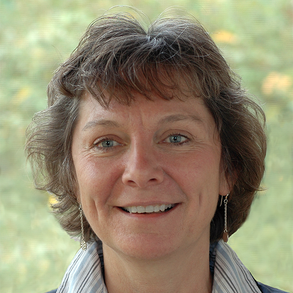 Author photo, portrait of Terese Allen framed by a blurry nature background, smiling slightly with wavy short cropped brown hair.