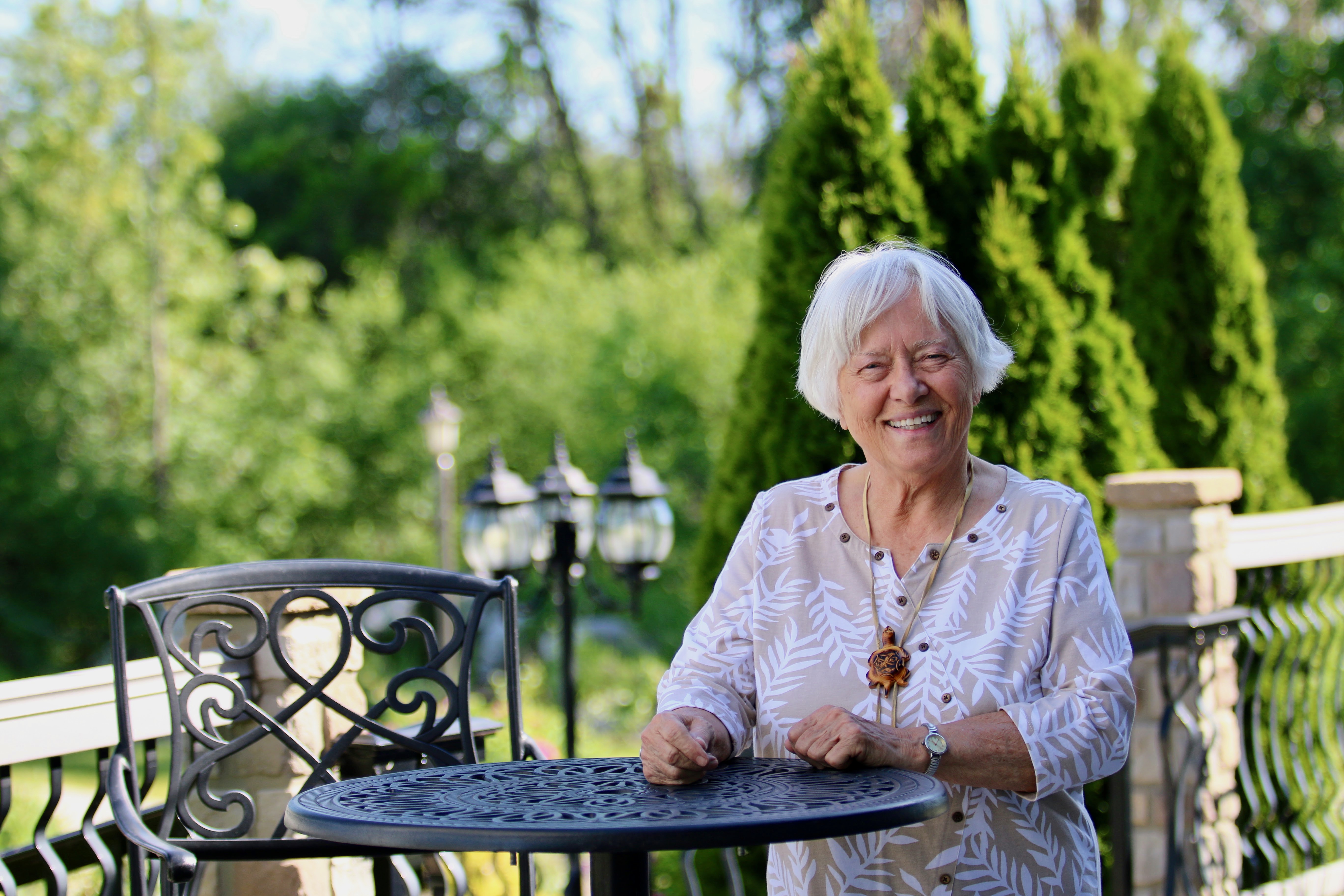 Mary Hilfiker sits at a table outside in the sunshine smiling at the camera. She has a white bob and is surrounded by flowers.