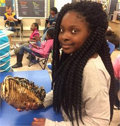 A young black girl with dreads holds a sea shell and smiles at the camera.