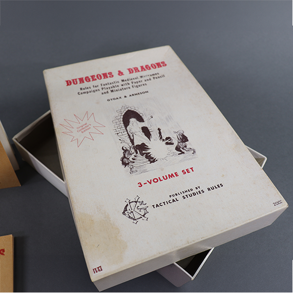 The white box that contained all the pieces of the set. It has red and black coloring for the text and features an illustration of a wizard fighting some sort of goblin-like monsters. It reads 'Dungeons and Dragons: Rules for fantastic medieval wargames campaigns playable with paper and pencil and miniature figures. Gygax & Arneson. 3-Volume Set. Published by Tactical Studies Rules.'