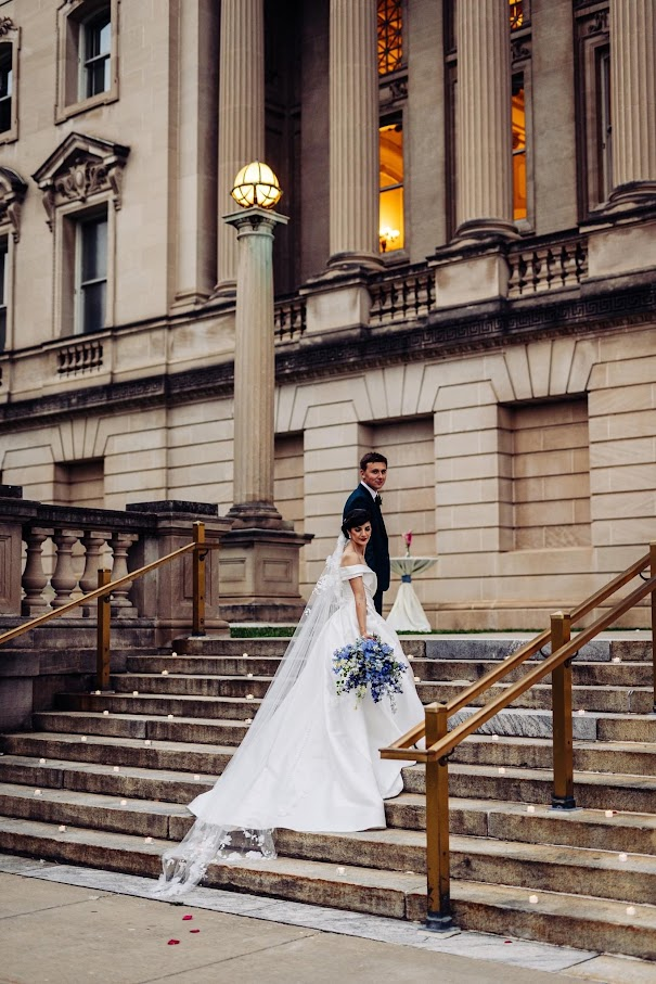 A bride and groom look back at the camera as they walk into the historic headquarters building. Her long dress flows over the steps.