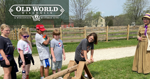 Old World Wisconsin. A group of children work with wooden device while being overseen by a woman in period clothing instructing them.