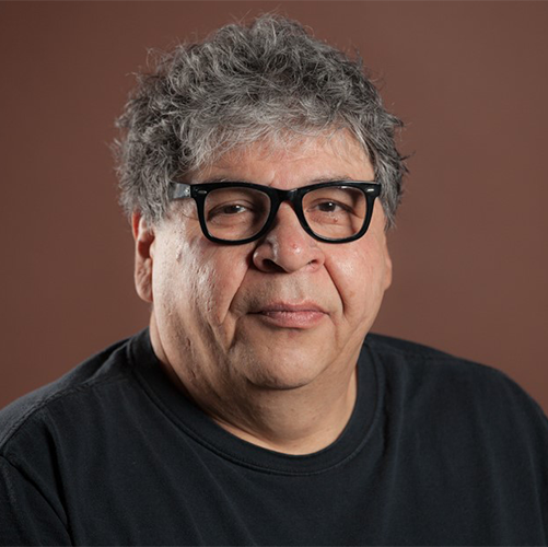 Author photo, portrait of Tom Weso on a rust background wearing a black shirt and black framed glasses. He's looking seriously at the camera.