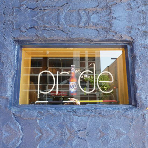 A neon sign with a bottle of Miller Lite for the 'i' spells “Pride” in the window of the 'Chances R' gay bar on Jay Street. The word 'Pride' is underlined with a neon rainbow. The window is framed in wood and surrounded by stucco painted blue.
