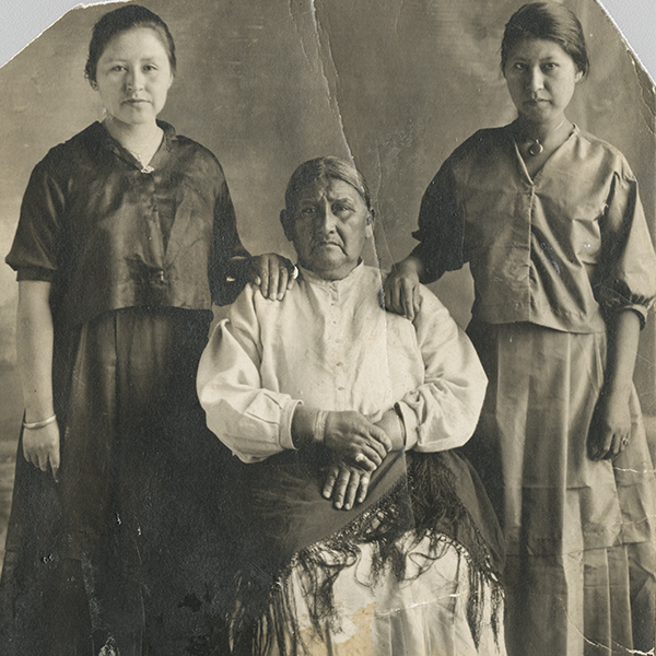 Full-length portrait of two Ho-Chunk women standing on either side of an older Ho-Chunk woman who is sitting. Each woman is wearing a long skirt, a blouse, and jewelry, and the woman sitting also has a shawl on her lap. Both standing women are resting a hand on the seated woman's shoulders.