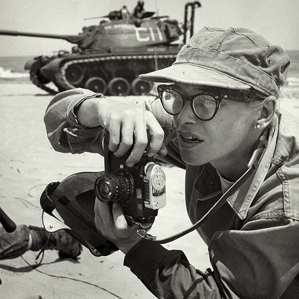 Dickey Chapelle, photographer, on the same Milwaukee beach where she learned to swim as a young girl. She was covering 'Operation Inland Seas' celebrating the opening of the St. Lawrence Seaway. She is holding her camera and there is a tank in the background. This is her favorite photograph of herself at work.