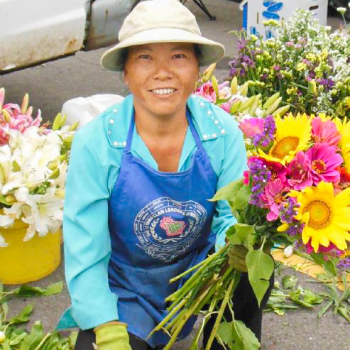 Hmong gardener kneeling and looking into the camera at the Northside Farmer’s Market. She is wearing a white hat, light blue shirt, royal blue apron, green gloves, black pants and black sandals. On the apron is the logo for 'The 12 Yang Clan Leaders of Wisconsin, Inc.' In her left hand is a bouquet of flowers, and flowers in buckets surround her.