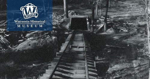 Wisconsin Historical Museum. A black and white photo shows an old mine hole leading under ground via train tracks, surrounded by felled trees.