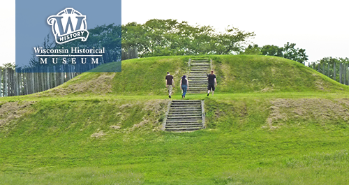 Wisconsin Historical Museum. A grassy hill that seems to be man made with stairs going up the middle, three modern day figures walk up the hill exploring ancient city structure of Aztalan.