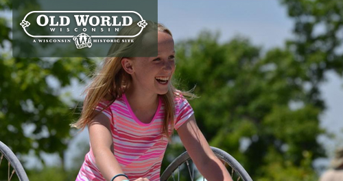 Old World Wisconsin. A blonde girl sits on a tall old fashioned bicycle wearing a pink, red, and white striped shirt.