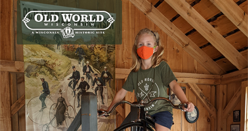Old World Wisconsin. A blonde girl sits on a tall old fashioned bicycle wearing a bright orange mask and an olive green shirt.