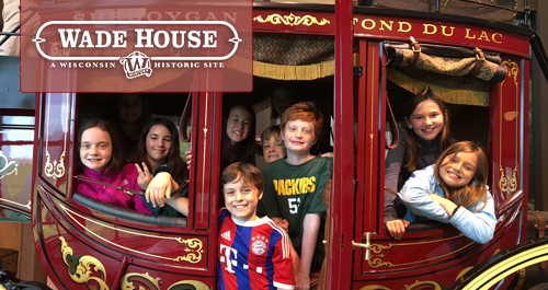 Wisconsin-Historical Museum. A large group of students and chaperones wearing matching shirts animatedly engage with a speaker off camera. Several girls raise their hands high!