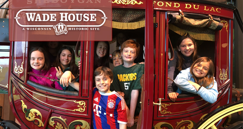Wisconsin-Historical Museum. A large group of students and chaperones wearing matching shirts animatedly engage with a speaker off camera. Several girls raise their hands high!