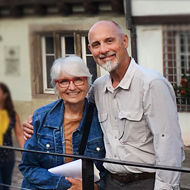 Pat and Maj. Gen. (Ret.) Al Wilkeni Sherry & John Stilin standing behind a black fence in front of a historic building. The streets are cobbled, the building is white stucko and wood beams. John has his arm around Sherry lovingly, she leans into him and they both smile happily at the camera. She's wearing a tan shirt wiht a denim jacket, her hair is white and cut short around her ears with swooping bangs, and red glasses. He is graying with a white goatee and a pale grey button up outdoors shirt and dark grey pants.