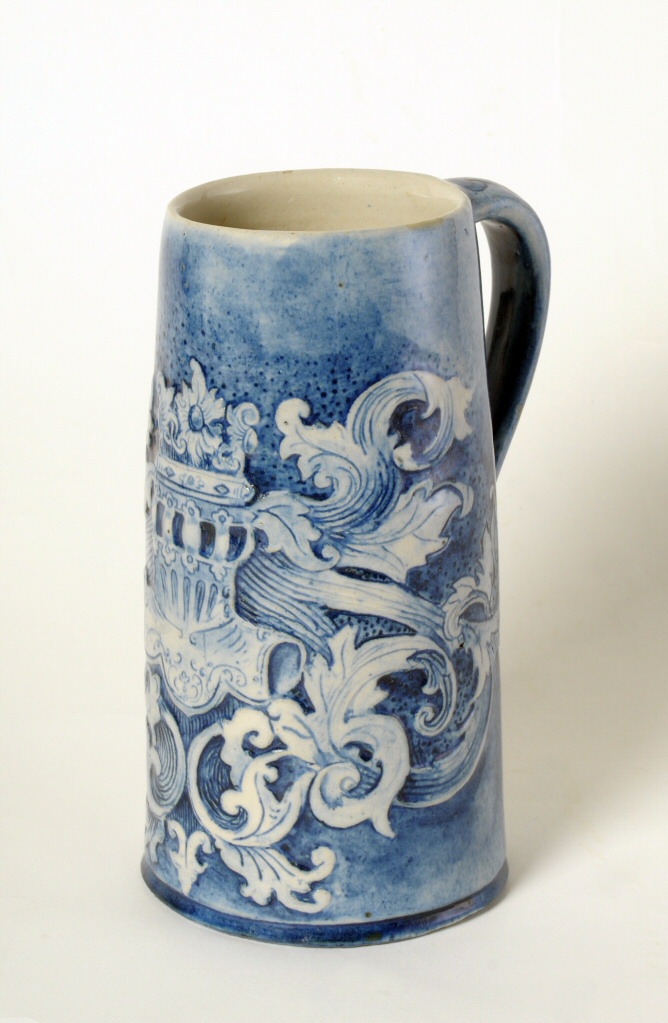 Art pottery stein, stoneware, blue, armor helmet motif and scroll design.